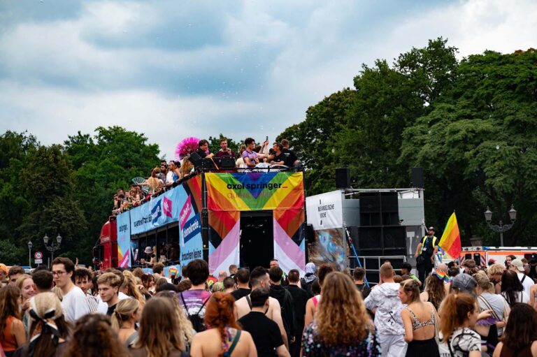 PACE events Eine große Menschenmenge versammelt sich im Freien zu einer Parade oder einem Festival. Die Menschen stehen auf und um einen mit Bannern, Regenbogenfarben und Flaggen dekorierten Festwagen, umgeben von Bäumen und unter einem bewölkten Himmel.