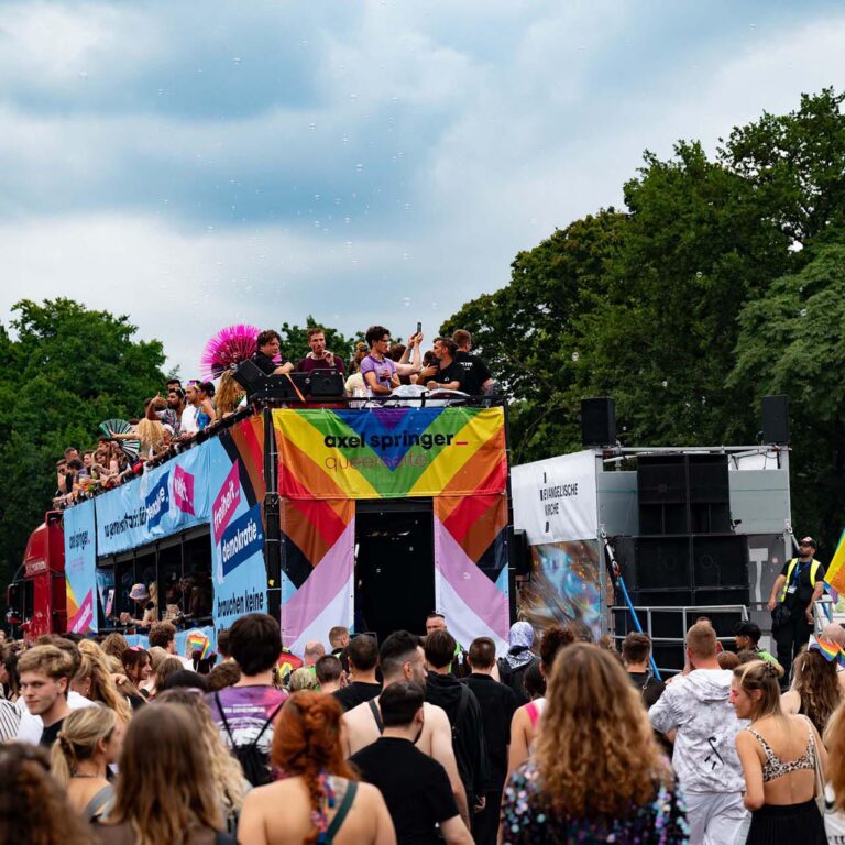 PACE events Eine große Menschenmenge versammelt sich im Freien zu einer Parade oder einem Festival. Die Menschen stehen auf und um einen mit Bannern, Regenbogenfarben und Flaggen dekorierten Festwagen, umgeben von Bäumen und unter einem bewölkten Himmel.