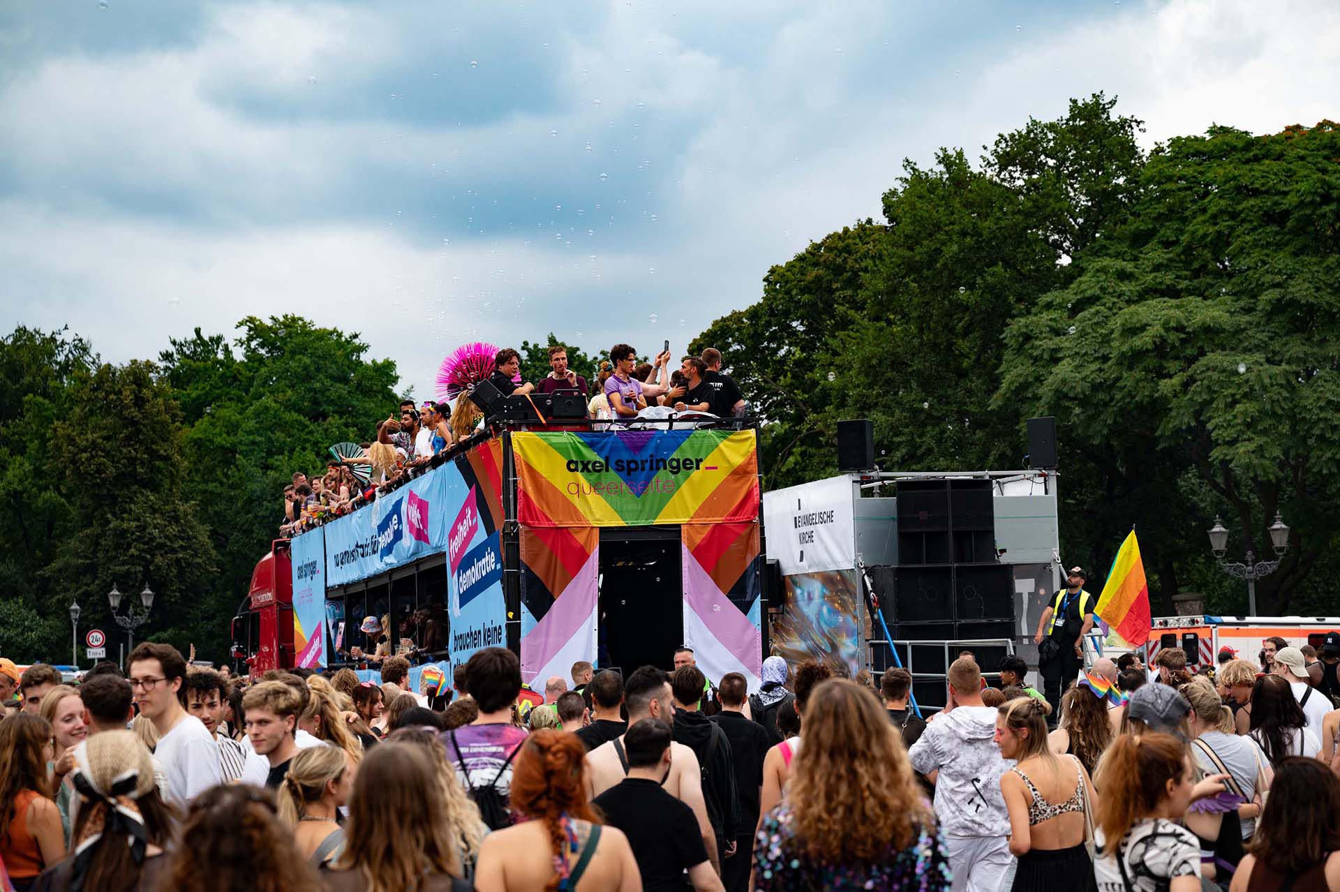 PACE events A large crowd gathers outdoors for a parade or festival. People stand on and around a float decorated with banners, rainbow colors and flags, surrounded by trees and under a cloudy sky.