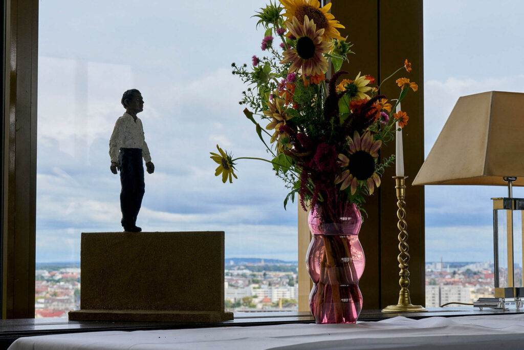 PACE events Eine kleine Statue, ein Strauß bunter Blumen in einer rosa Vase, eine Lampe und eine Kerze stehen auf einem Tisch neben einem großen Fenster mit Blick auf eine Stadtlandschaft unter einem bewölkten Himmel.