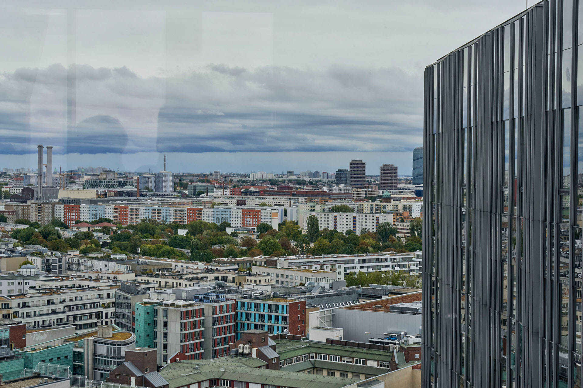PACE events Eine Stadtansicht mit mittelhohen Gebäuden, grünen Bäumen und einem bewölkten Himmel, mit einem modernen Glasgebäude auf der rechten Seite, das einen Teil der Stadt widerspiegelt.