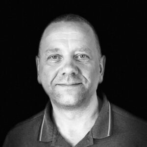 PACE events Ein Mann mittleren Alters mit kurzen Haaren lächelt sanft in die Kamera. Er trägt ein Hemd mit Kragen und der Hintergrund ist einfarbig schwarz, so dass sein Gesicht im Mittelpunkt des Schwarz-Weiß-Bildes steht.