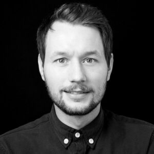PACE events Ein Mann mit kurzen dunklen Haaren und einem Bart, der ein zugeknöpftes dunkles Hemd trägt, blickt in die Kamera und lächelt leicht vor einem schlichten schwarzen Hintergrund.
