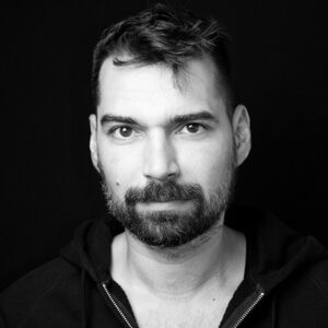 PACE events Ein Mann mit kurzen dunklen Haaren und einem Bart blickt mit neutralem Gesichtsausdruck direkt in die Kamera, vor einem schlichten schwarzen Hintergrund. Er trägt ein dunkles Oberteil mit Reißverschluss. Das Foto ist in schwarz-weiß gehalten.
