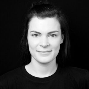 PACE events Eine Frau mit glattem, nach hinten gezogenem dunklem Haar blickt in die Kamera und lächelt leicht. Sie trägt ein schlichtes dunkles Hemd und steht vor einem schwarzen Hintergrund. Das Bild ist in Schwarz-Weiß gehalten.
