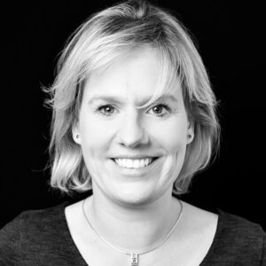 PACE events Eine Frau mit kurzem, hellblondem Haar lächelt in die Kamera. Sie trägt ein dunkles Oberteil, kleine Ohrringe und eine Halskette mit einem rechteckigen Anhänger. Der Hintergrund ist schwarz. Das Bild ist in schwarz-weiß gehalten.