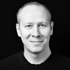PACE events Schwarz-Weiß-Porträt eines Mannes mit kurzen hellen Haaren, der einen dunklen gerippten Pullover trägt, leicht lächelt und direkt in die Kamera blickt, vor einem schlichten dunklen Hintergrund.
