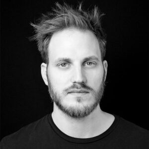 PACE events Ein Mann mit heller Haut, kurzem, zerzaustem Haar und Bart blickt mit neutralem Blick direkt in die Kamera. Er trägt ein schlichtes dunkles T-Shirt vor einem schwarzen Hintergrund.