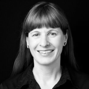 PACE events Eine Frau mit glattem, hellbraunem Haar und Pony lächelt in die Kamera. Sie trägt ein dunkles Hemd und kleine runde Ohrringe, und der Hintergrund ist einfarbig schwarz.