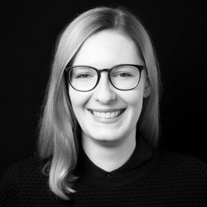 PACE events Eine Frau mit glattem, schulterlangem hellem Haar und Brille lächelt in die Kamera. Sie trägt einen strukturierten dunklen Pullover und ein Hemd mit Kragen und posiert vor einem schlichten schwarzen Hintergrund.