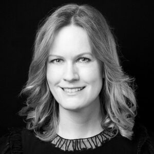 PACE events Ein Schwarz-Weiß-Porträt einer Frau mit schulterlangem, gewelltem Haar, die sanft lächelt und ein dunkles Oberteil mit durchsichtigem Ausschnitt vor einem schlichten schwarzen Hintergrund trägt.