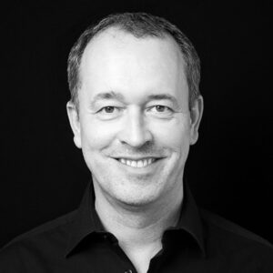 PACE events Ein Mann mittleren Alters mit kurzen Haaren und einem dunklen Hemd mit Kragen lächelt in einem Schwarz-Weiß-Porträt vor einem schlichten schwarzen Hintergrund in die Kamera.