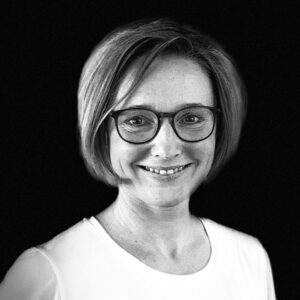 PACE events Schwarz-Weiß-Porträt einer lächelnden Frau mit kurzen, hellen Haaren, Brille und weißem Oberteil vor einem schlichten dunklen Hintergrund.