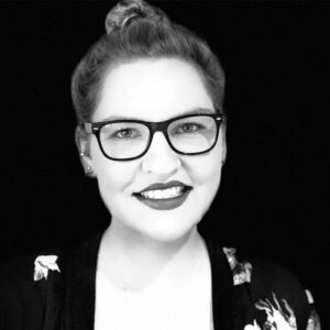 PACE events Schwarz-Weiß-Porträt einer lächelnden Frau mit hellem Haar in einem Dutt, die eine dunkel umrandete Brille, Lippenstift und ein gemustertes Oberteil trägt, vor einem schlichten schwarzen Hintergrund.