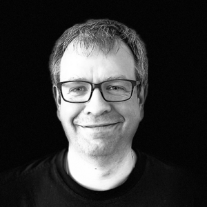 PACE events Schwarz-Weiß-Porträt einer Person mittleren Alters mit kurzen Haaren, Brille und einem leichten Lächeln, die ein dunkles Hemd vor einem schlichten schwarzen Hintergrund trägt.