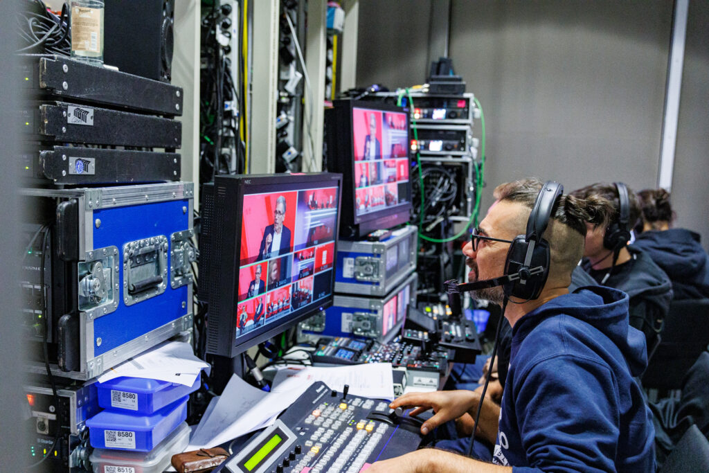 PACE events Ein Mann mit einem Headset bedient die Videoproduktionsausrüstung in einem Kontrollraum und überwacht mehrere Bildschirme, die eine Live-Veranstaltung mit einem Sprecher auf der Bühne zeigen. Andere Crew-Mitglieder arbeiten in der Nähe inmitten von Kabeln und technischer Ausrüstung.
