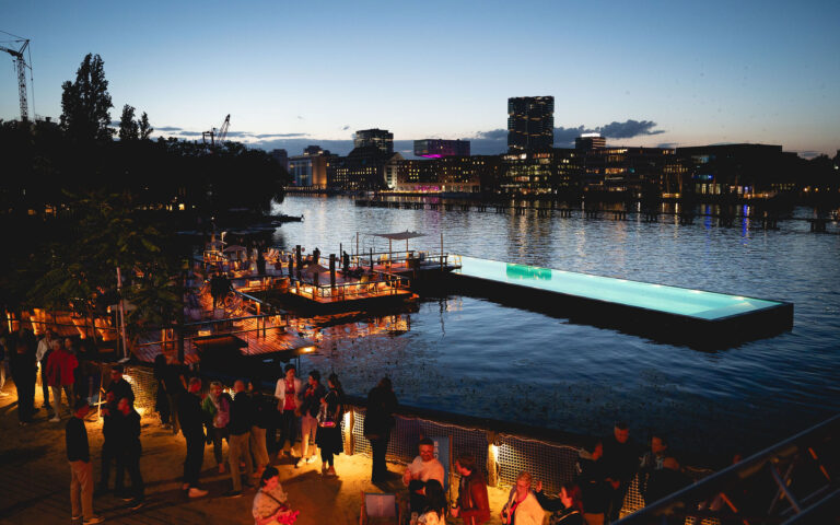 PACE events Menschen versammeln sich in der Abenddämmerung an einem hell erleuchteten Teich am Flussufer, wobei die Gebäude der Stadt und ihre Spiegelungen auf dem Wasser unter dem sich verdunkelnden Himmel sichtbar sind.