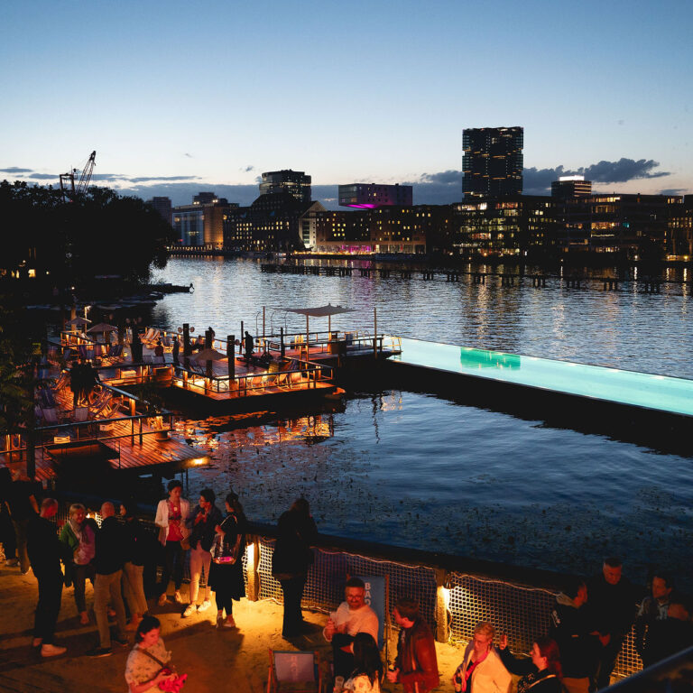 PACE events Menschen versammeln sich in der Abenddämmerung an einem hell erleuchteten Teich am Flussufer, wobei die Gebäude der Stadt und ihre Spiegelungen auf dem Wasser unter dem sich verdunkelnden Himmel sichtbar sind.
