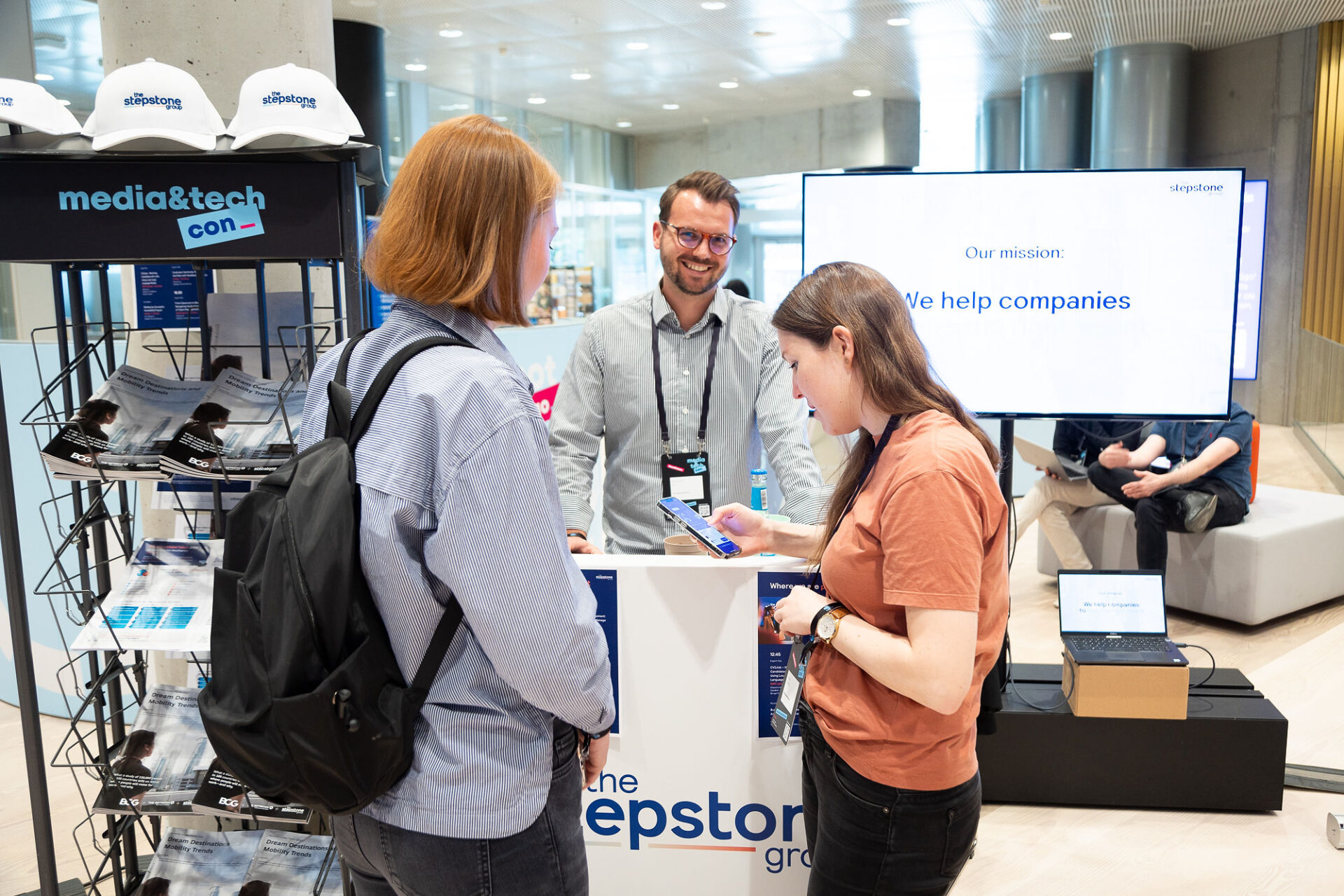 PACE events Drei Personen unterhalten sich an einem Stand auf einer Technologiekonferenz mit Broschüren, Markenhüten und einem großen Bildschirm mit der Aufschrift "Unsere Mission: Wir helfen Unternehmen." Eine Person steht hinter dem Stand und lächelt zwei Teilnehmerinnen an.