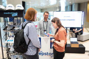 PACE events Drei Personen unterhalten sich an einem Stand auf einer Technologiekonferenz mit Broschüren, Markenhüten und einem großen Bildschirm mit der Aufschrift "Unsere Mission: Wir helfen Unternehmen." Eine Person steht hinter dem Stand und lächelt zwei Teilnehmerinnen an.
