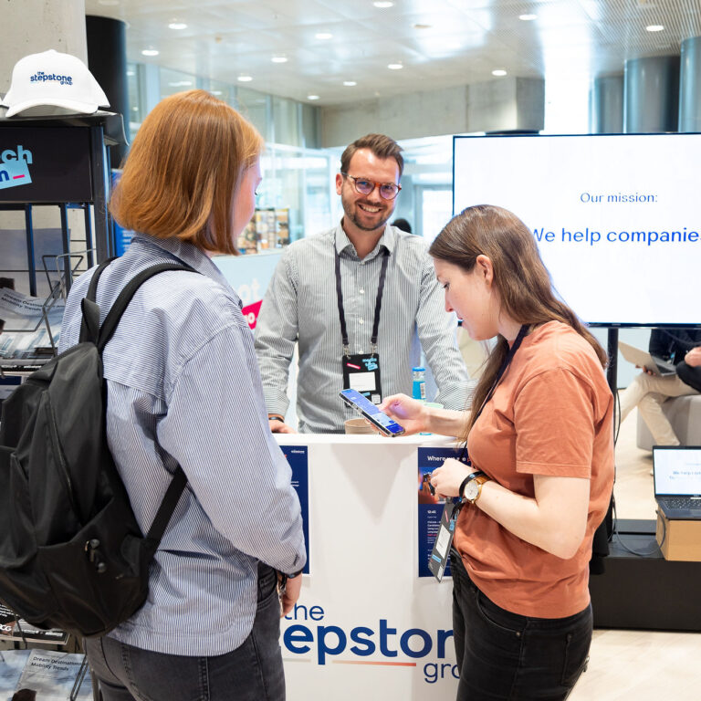 PACE events Drei Personen unterhalten sich an einem Stand auf einer Technologiekonferenz mit Broschüren, Markenhüten und einem großen Bildschirm mit der Aufschrift "Unsere Mission: Wir helfen Unternehmen." Eine Person steht hinter dem Stand und lächelt zwei Teilnehmerinnen an.