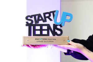 PACE events Eine Person hält eine Trophäe mit der Aufschrift STARTUP TEENS. Auf dem Sockel steht STARTUP TEENS FINALE 2024 1. Platz Bildung. Die Buchstaben der Trophäe sind schwarz und blau, und die Person hat lackierte Nägel und ein Tattoo am Handgelenk.