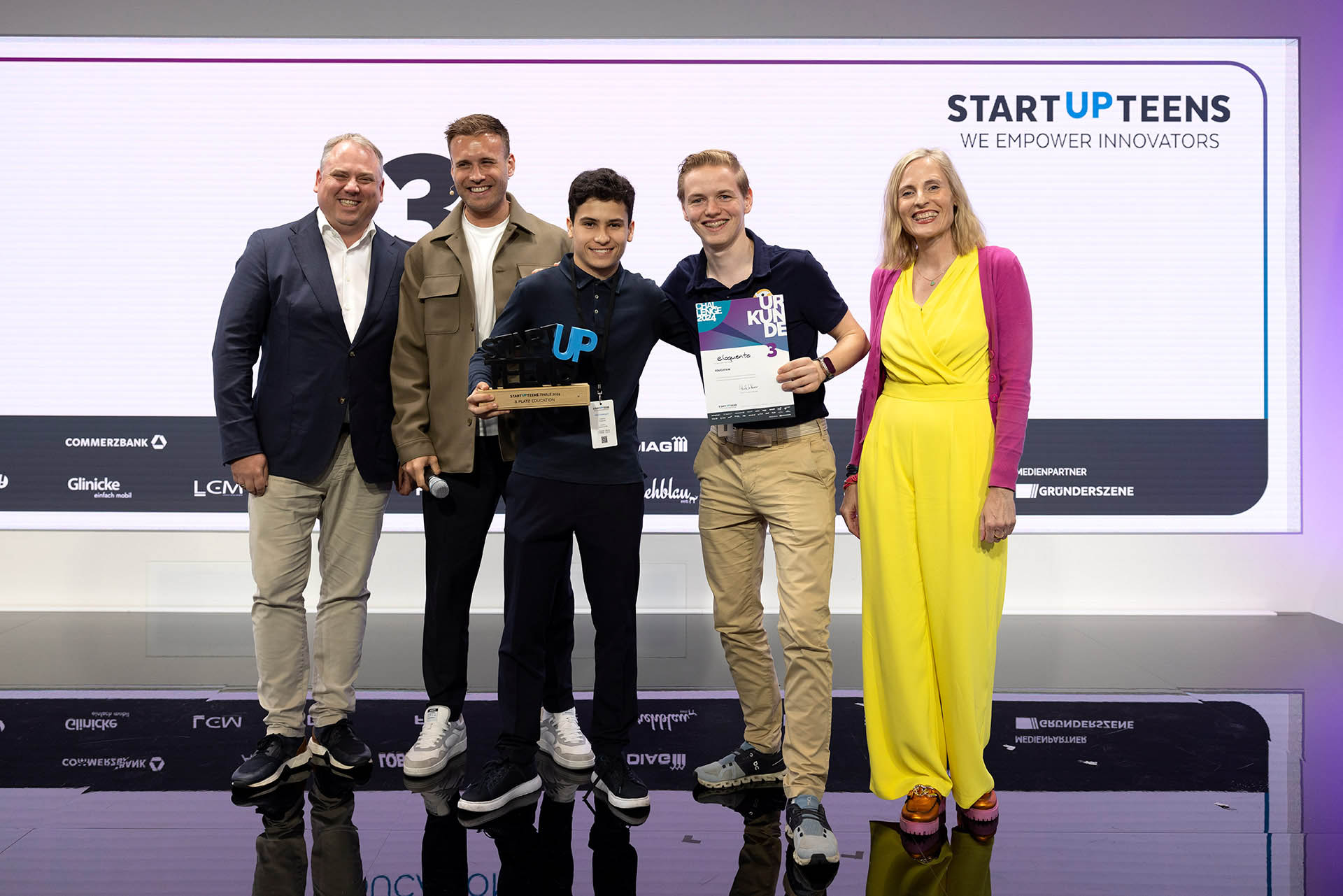 PACE events Fünf Personen stehen auf einer Bühne bei einer STARTUPTEENS-Veranstaltung. Zwei junge Männer in der Mitte halten eine Trophäe und eine Urkunde, während drei Erwachsene lächelnd daneben stehen. Hinter ihnen ist ein großes STARTUPTEENS-Schild zu sehen.
