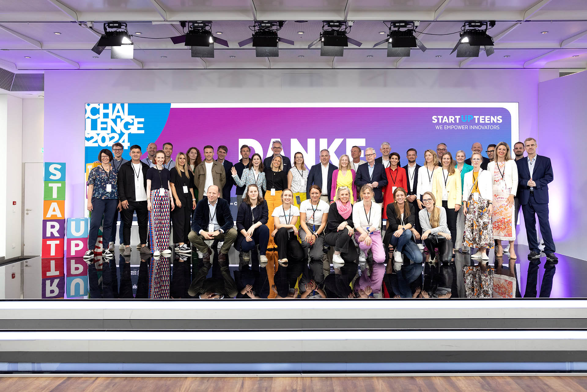 PACE events Eine große Gruppe von Menschen posiert auf einer Bühne mit einem bunten Hintergrund, auf dem CHALLENGE 2024, DANKE und STARTUPTEENS zu sehen sind. Die Atmosphäre wirkt förmlich, und die Gruppe besteht aus verschiedenen Altersgruppen und Geschlechtern.