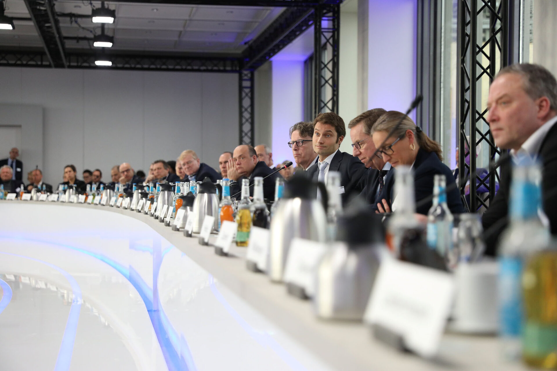 PACE events Eine Gruppe von formell gekleideten Personen sitzt um einen langen, geschwungenen Konferenztisch mit Mikrofonen, Namensschildern, Wasserflaschen und Kaffeekannen und nimmt an einer geschäftlichen Besprechung in einem modernen, gut beleuchteten Raum teil.