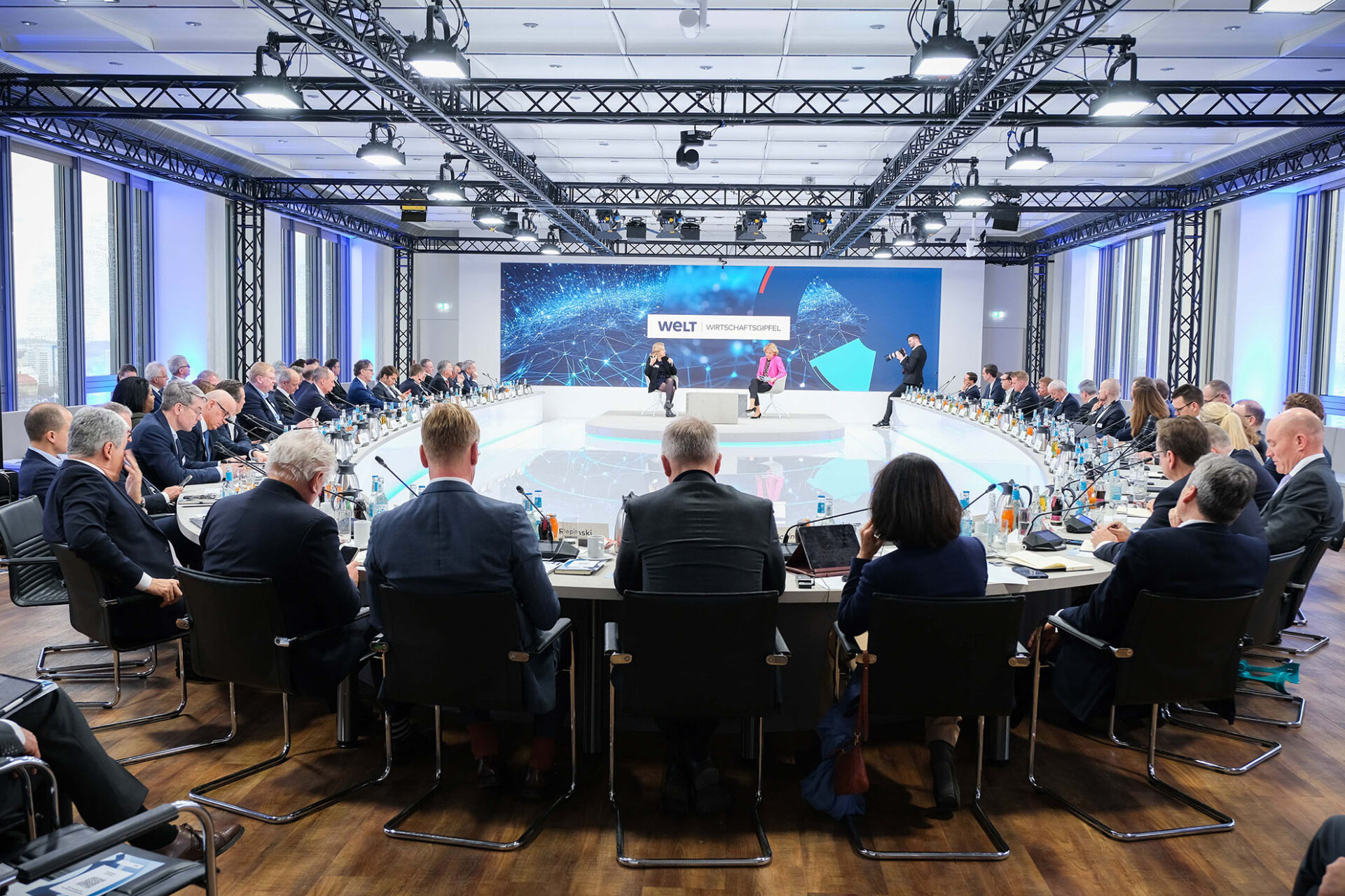 PACE events Ein großer Konferenzraum mit vielen Menschen in Anzügen, die um einen runden Tisch sitzen und auf eine Bühne blicken, auf der Redner vor einem großen Bildschirm mit der Aufschrift "WELT Wirtschaftsgipfel" präsentieren.