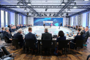 PACE events Ein großer Konferenzraum mit vielen Menschen in Anzügen, die um einen runden Tisch sitzen und auf eine Bühne blicken, auf der Redner vor einem großen Bildschirm mit der Aufschrift "WELT Wirtschaftsgipfel" präsentieren.