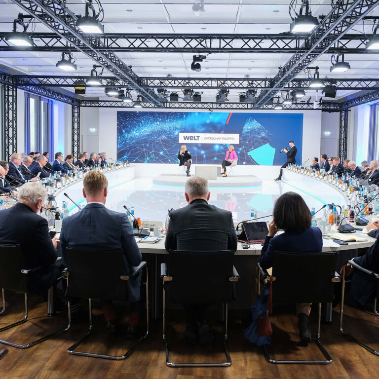 PACE events Ein großer Konferenzraum mit vielen Menschen in Anzügen, die um einen runden Tisch sitzen und auf eine Bühne blicken, auf der Redner vor einem großen Bildschirm mit der Aufschrift "WELT Wirtschaftsgipfel" präsentieren.