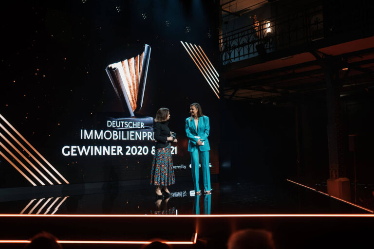 PACE events Zwei Frauen stehen bei einer Preisverleihung auf der Bühne. Hinter ihnen zeigt eine große Leinwand den Text Deutscher Immobilienpreis Gewinner 2020 & 2021 mit einer Trophäengrafik. Die Bühne ist dunkel mit leuchtenden orangefarbenen Linien.