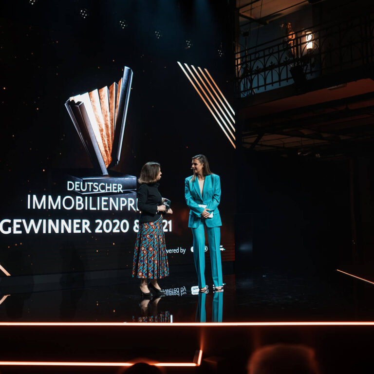 PACE events Zwei Frauen stehen bei einer Preisverleihung auf der Bühne. Hinter ihnen zeigt eine große Leinwand den Text Deutscher Immobilienpreis Gewinner 2020 & 2021 mit einer Trophäengrafik. Die Bühne ist dunkel mit leuchtenden orangefarbenen Linien.