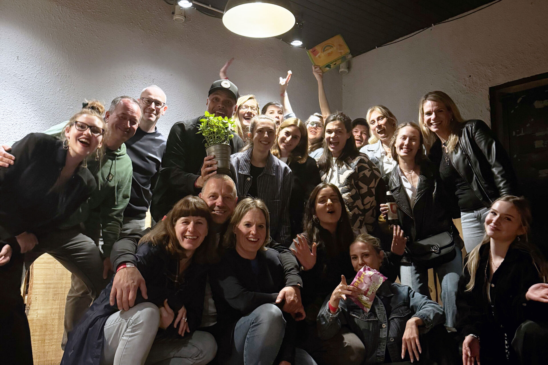PACE events Eine große Gruppe von Menschen posiert in einem Innenraum unter einer Deckenleuchte, einige kniend, andere stehend, lächelnd und winkend in die Kamera. Eine Person hält eine Topfpflanze, eine andere eine Tüte mit Süßigkeiten.