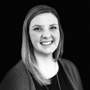 PACE events Eine junge Frau mit glattem, hellem Haar lächelt in die Kamera. Sie trägt ein dunkles Oberteil und eine strukturierte Strickjacke. Der Hintergrund ist komplett schwarz, wodurch sich ihr Gesicht und ihr Oberkörper abheben. Das Foto ist in Schwarz-Weiß gehalten.