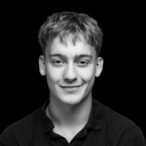 PACE events Schwarz-Weiß-Porträt eines jungen Mannes mit kurzem, zerzaustem Haar, der ein dunkles Hemd mit Kragen trägt und leicht lächelt, während er direkt in die Kamera schaut, vor einem schlichten schwarzen Hintergrund.