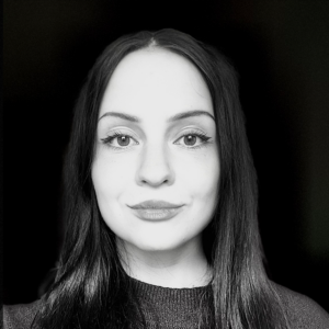 PACE events Eine junge Frau mit langen dunklen Haaren und heller Haut schaut mit neutralem Ausdruck direkt in die Kamera. Das Foto ist schwarz-weiß, mit dunklem Hintergrund und weicher Beleuchtung auf ihrem Gesicht.