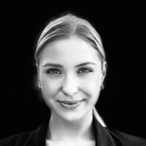 PACE events Schwarzes und weißes Nahaufnahmeporträt einer jungen Frau mit hellem, zurückgestecktem Haar, die Ohrringe und einen dunklen Blazer trägt und vor einem schwarzen Hintergrund leicht lächelt.