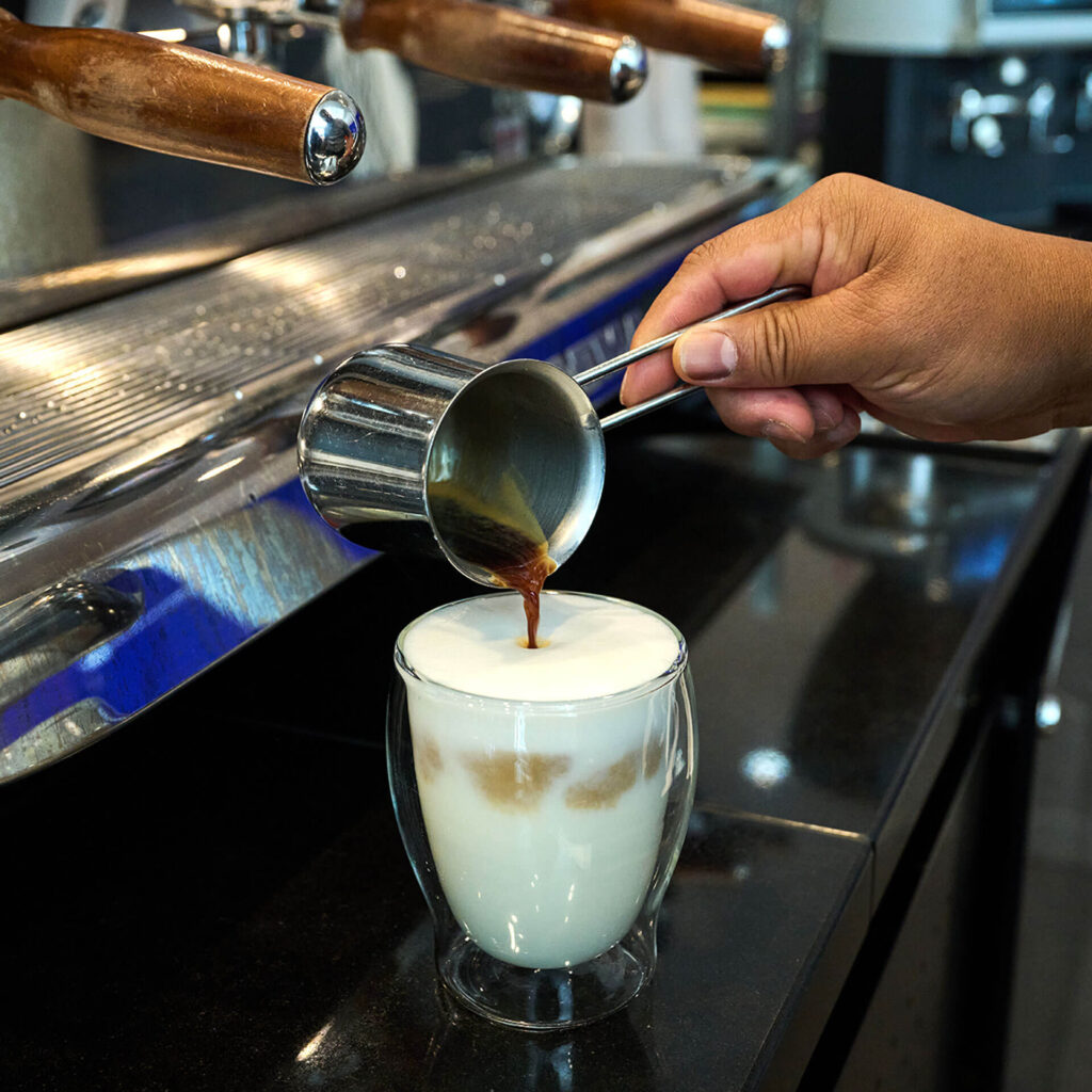 PACE events Eine Hand gießt Espresso aus einer kleinen Metallkanne in ein Glas mit schaumiger Milch, wodurch ein Milchkaffee entsteht. Die Szene ist auf einer schwarzen Arbeitsplatte neben einer Espressomaschine aufgebaut.