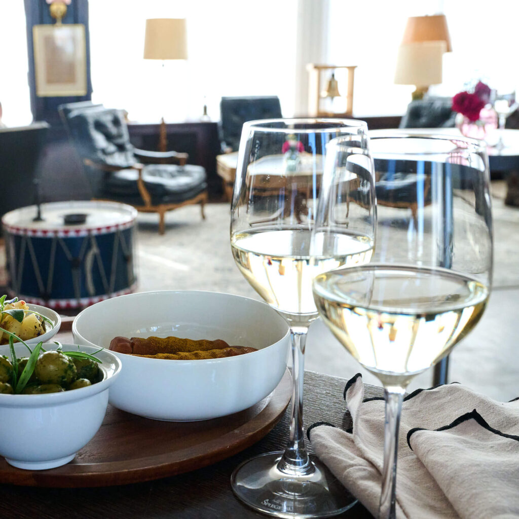 PACE events Zwei Gläser Weißwein, eine Schüssel mit Salat und eine Schüssel mit Essen stehen auf einem Holztablett auf einem Tisch, während im unscharfen Hintergrund ein gemütliches Wohnzimmer mit Sesseln und Trommeln zu sehen ist.