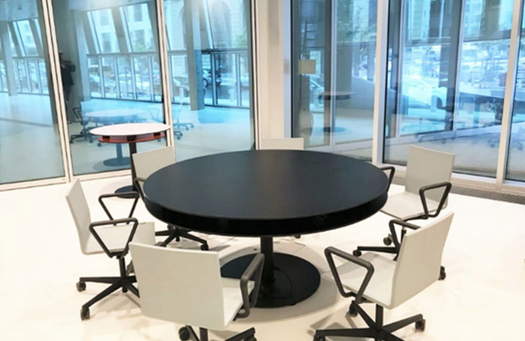 PACE events Ein moderner Besprechungsraum mit einem runden schwarzen Tisch, um den sechs weiße Bürostühle auf Rollen stehen. Große Glasfenster und -türen lassen Tageslicht herein und bieten einen Blick auf die städtische Umgebung. Im Hintergrund steht ein kleiner runder weißer Tisch.