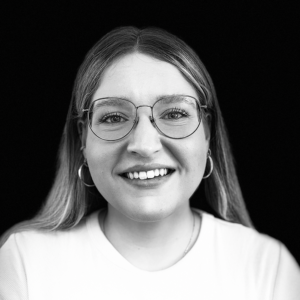 PACE events Eine Person mit langen Haaren, Brille und Creolen lächelt in die Kamera. Sie trägt ein helles Hemd, und der Hintergrund ist komplett schwarz. Das Bild ist in Schwarz-Weiß gehalten.