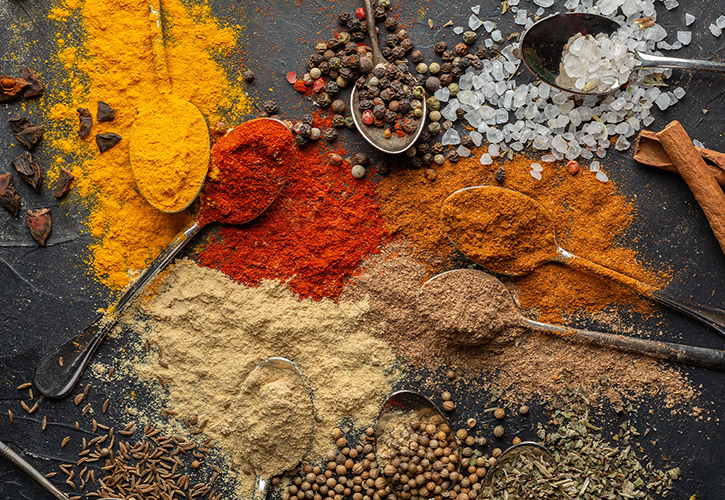 Verschiedene bunte Gewürze und Kräuter, darunter gemahlene Pulver und ganze Samen, werden mit Metalllöffeln kunstvoll auf einer dunklen Fläche arrangiert. Zu den sichtbaren Gewürzen gehören Kurkuma, Paprika, Salz, Pfefferkörner und getrocknete Kräuter.