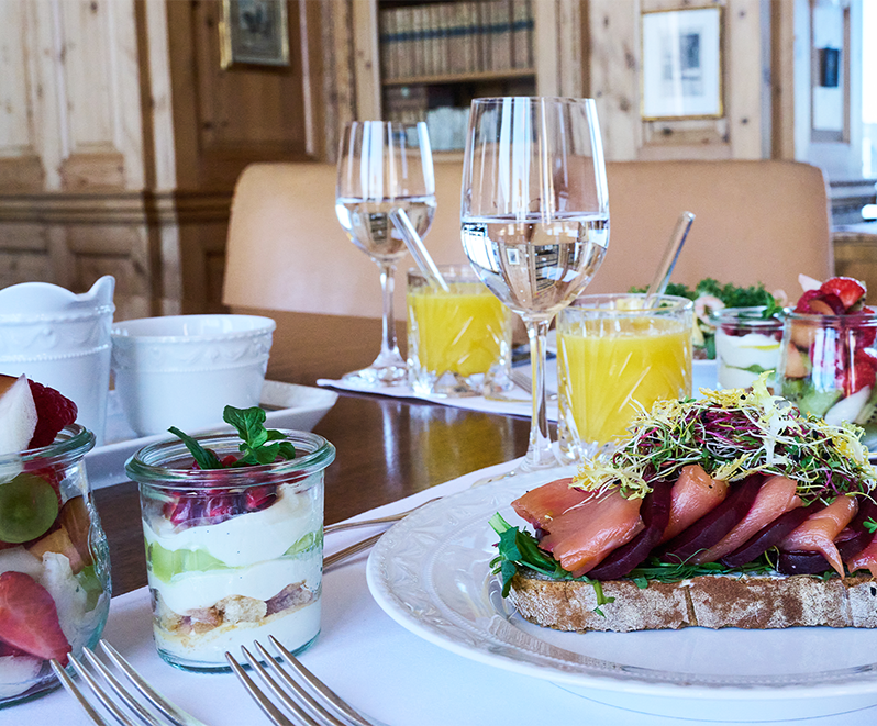 Ein elegantes Frühstück auf einem Tisch mit geräuchertem Lachs auf Brot mit Grünzeug, Parfaits mit Obst und Joghurt in Gläsern, Wasser und Orangensaft in einem gemütlichen, holzgetäfelten Speisesaal.