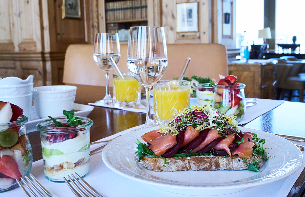 Ein elegantes Frühstück auf einem Tisch mit geräuchertem Lachs auf Brot mit Grünzeug, Parfaits mit Obst und Joghurt in Gläsern, Wasser und Orangensaft in einem gemütlichen, holzgetäfelten Speisesaal.