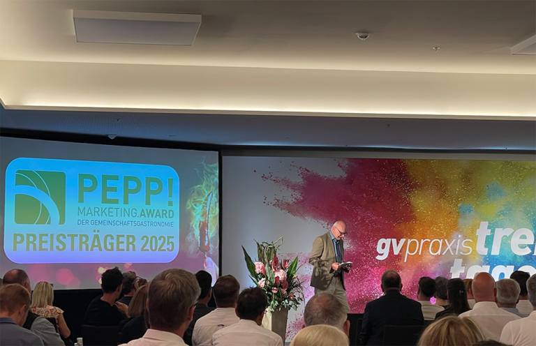 Ein Redner steht auf der Bühne der gv praxis-Veranstaltung und wendet sich an das Publikum. Auf einer Leinwand ist der PEPP! Marketing Award Preisträger 2025 neben bunten Wandbildern und einem Blumenarrangement.