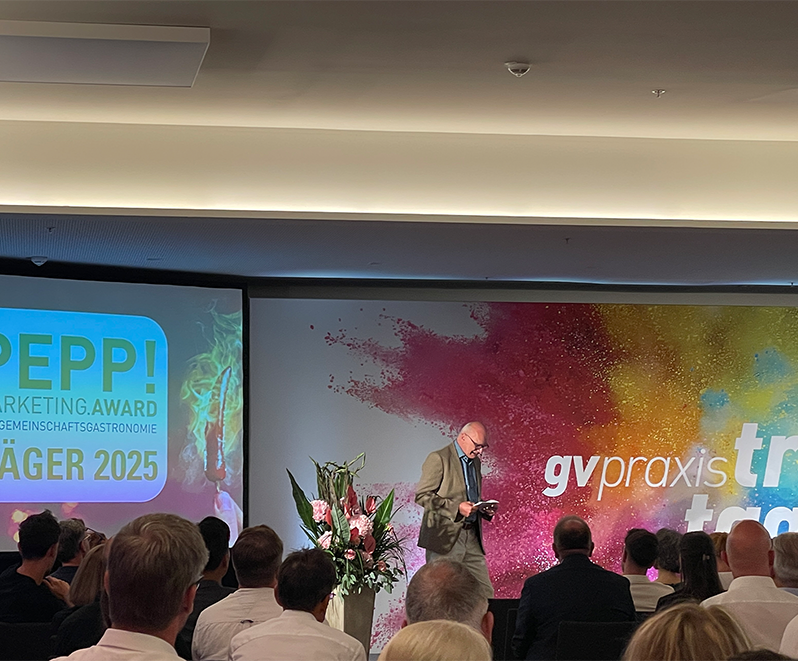 Ein Redner steht auf der Bühne der gv praxis-Veranstaltung und wendet sich an das Publikum. Auf einer Leinwand ist der PEPP! Marketing Award Preisträger 2025 neben bunten Wandbildern und einem Blumenarrangement.