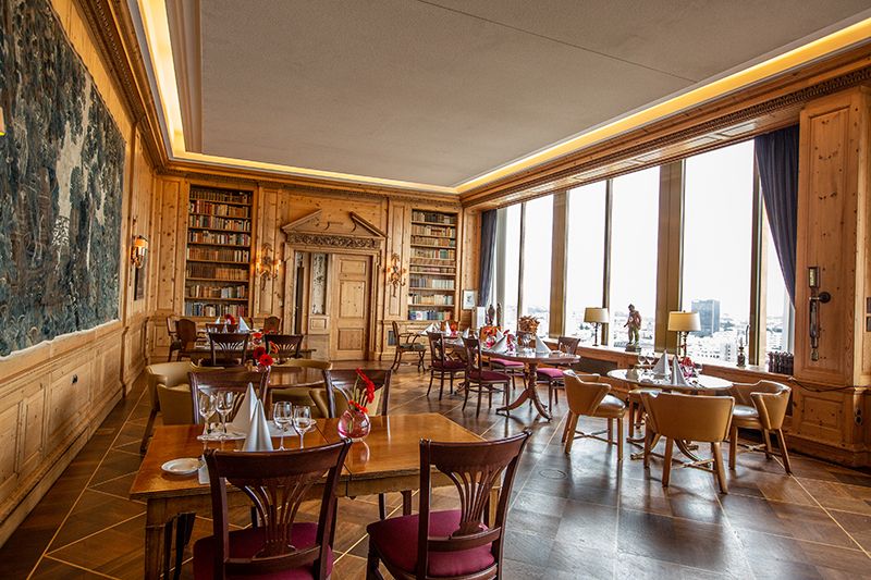Elegantes Restaurant mit Holzwänden, großen Fenstern mit Blick auf die Stadt, Bücherregalen, ordentlich gedeckten Tischen, gepolsterten Stühlen und natürlichem Sonnenlicht, das den gemütlichen, raffinierten Raum erhellt.