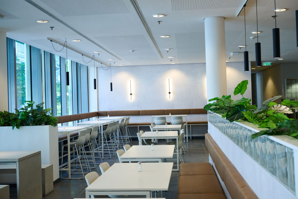A modern, empty cafeteria with white tables, brown and white chairs, indoor plants, large windows on the left-hand side and wall lights on a white wall. The room is brightly lit and clearly laid out.